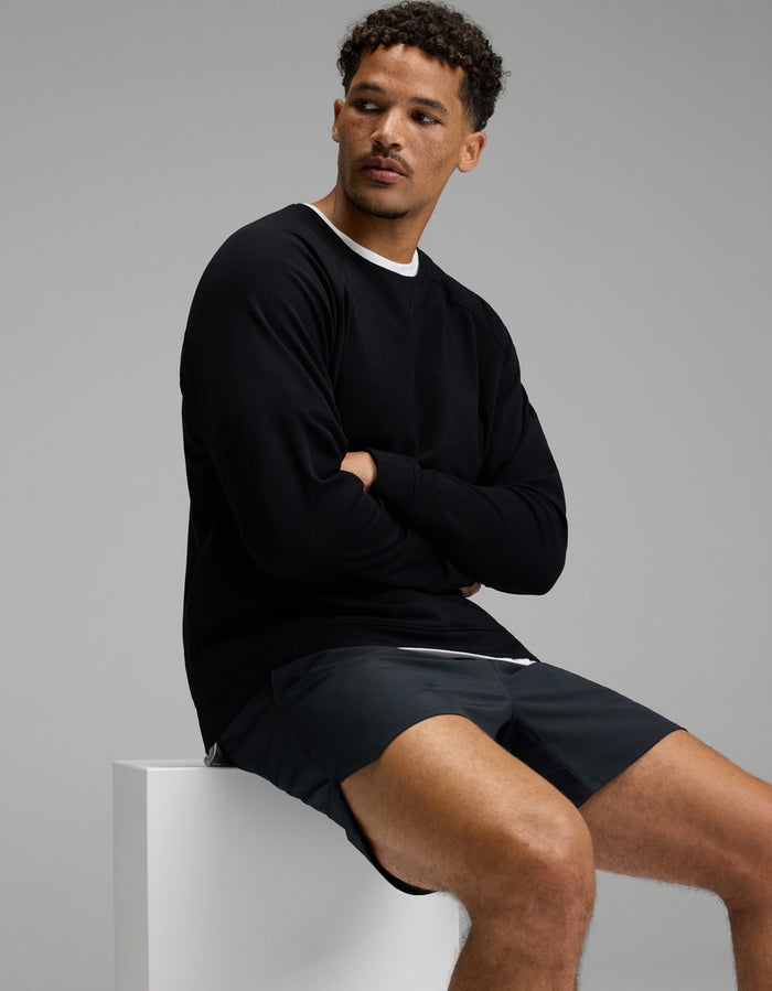 A man with short curly hair sits on a white block, wearing Cozy Earth’s Men’s Brushed Bamboo Pullover Crew in black over a white shirt and dark shorts, looking to his left against a plain gray background.|Color:Black