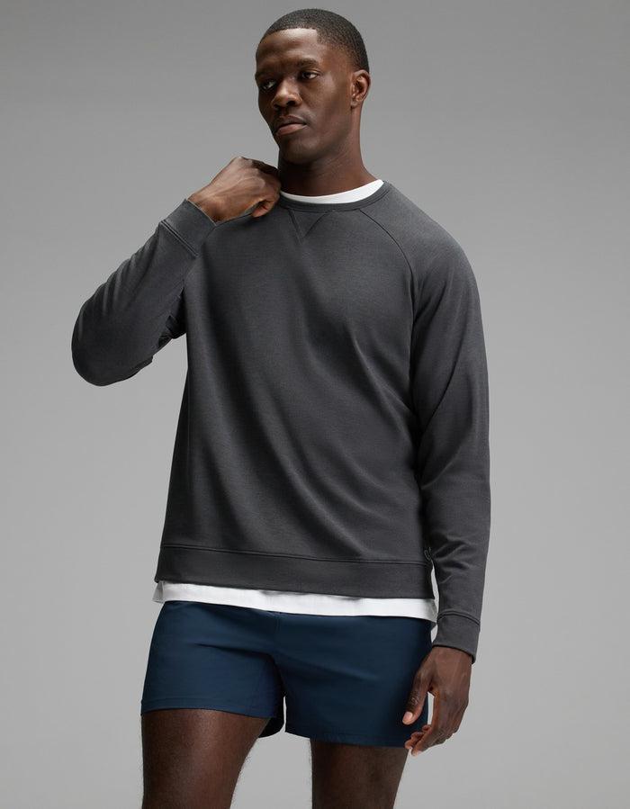 A man wearing the Cozy Earth Men’s Brushed Bamboo Pullover Crew in Charcoal over a white shirt and navy blue shorts stands against a plain gray background, gazing slightly to the side with one hand near his neck.|Color:Charcoal