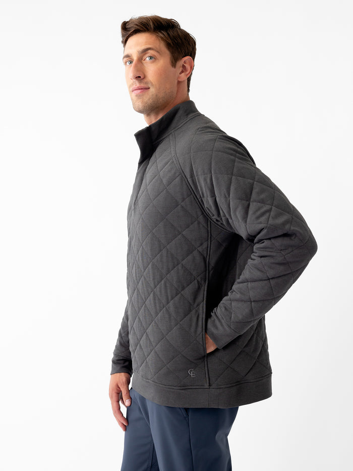 A man wearing the Men's Ultra-Soft Bamboo Quilted Snap Pullover in dark gray by Cozy Earth and dark blue pants stands against a plain white background. He is posing with both hands in his pockets and looking slightly to his left. The pullover features a high collar and a quilted pattern. |Color:Charcoal