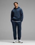 A person stands against a plain gray background, wearing the Cozy Earth Men’s Cityscape Hoodie & Sweatpant Set in Eclipse, paired with white sneakers and a white undershirt. Their hands are in their pockets and they are looking straight ahead.|Color:Eclipse