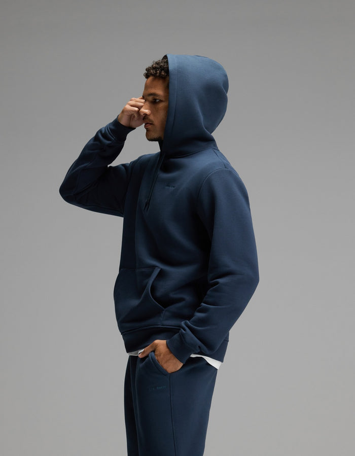 A person dressed in the Cozy Earth Men’s Cityscape Hoodie & Sweatpant Set in navy stands in profile against a plain gray background, with one hand touching their forehead.|Color:Eclipse