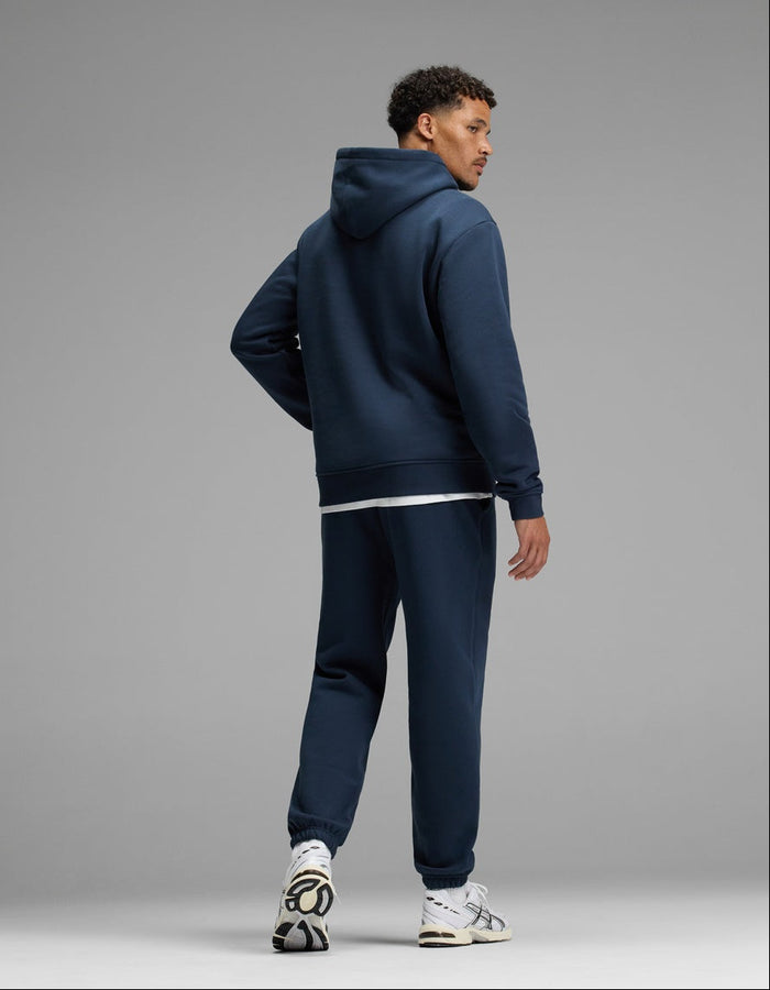 A man stands facing slightly away, wearing the Cozy Earth Men’s Cityscape Hoodie & Sweatpant Set in navy blue with white sneakers against a plain gray background.|Color:Eclipse
