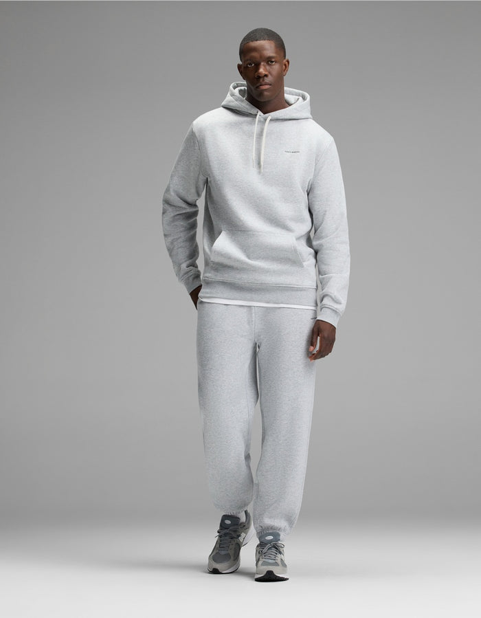 A man stands against a gray backdrop wearing the Cozy Earth Men’s Cityscape Hoodie & Sweatpant Set in Heather Grey, paired with gray athletic shoes. His left hand is in his pocket, and he faces forward with a neutral expression.|Color:Heather Grey