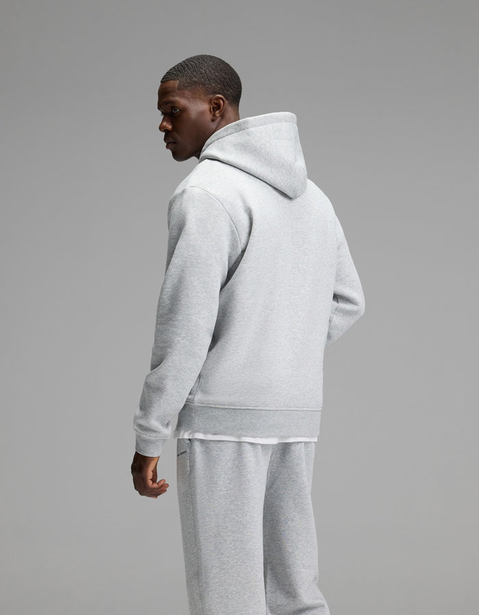 A man models the Cozy Earth Men’s Cityscape Hoodie & Sweatpant Set in light gray, standing against a plain gray background and glancing over his left shoulder.|Color:Heather Grey