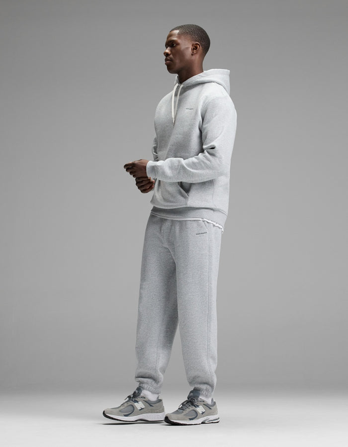 A person stands in profile against a gray background, wearing the Cozy Earth Men’s Cityscape Hoodie & Sweatpant Set paired with gray sneakers. The outfit looks casual and comfortable.|Color:Heather Grey