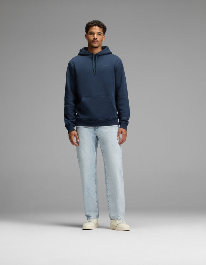 A man stands facing forward against a plain gray background, wearing the Cozy Earth Men’s Cityscape Hoodie in navy blue, paired with light blue jeans and off-white sneakers.|Color:Eclipse