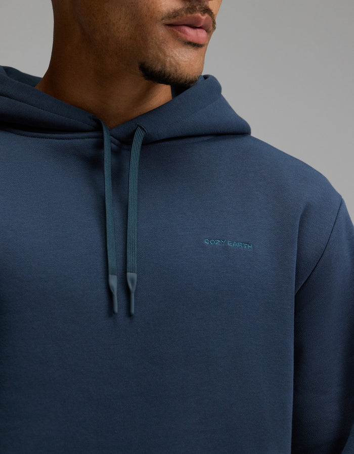 A person wearing the Men’s Cityscape Hoodie by Cozy Earth in dark blue, shown from the shoulders to the mouth, with “Cozy Earth” embroidered on the chest and hood strings hanging down.|Color:Eclipse