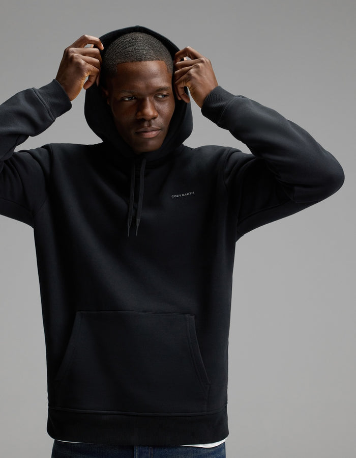 A man wears the Cozy Earth Men’s Cityscape Hoodie in Jet Black, lifting the hood over his head as he stands against a plain gray background, gazing to the side with a neutral expression.|Color:Jet Black