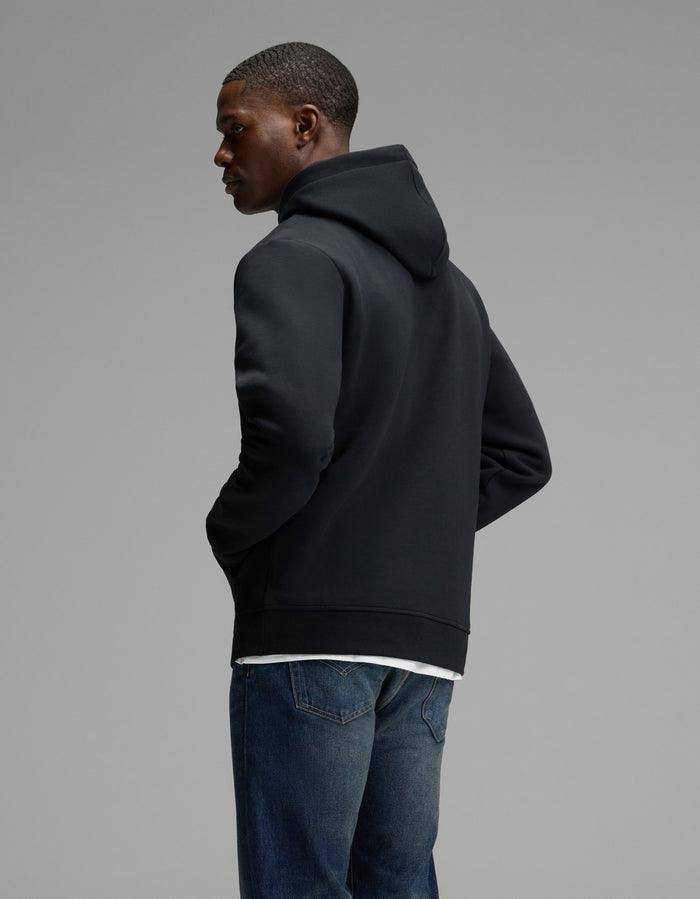 A person wearing the Cozy Earth Men’s Cityscape Hoodie in black and blue jeans stands with hands in pockets, facing away and glancing to the side against a plain gray background.|Color:Jet Black