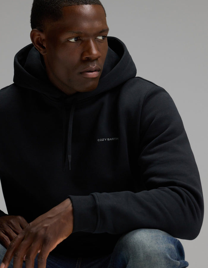A man wearing the Men’s Cityscape Hoodie by Cozy Earth, featuring the brand’s text on the front, looks to his left with one arm resting on his knee against a plain background.|Color:Jet Black