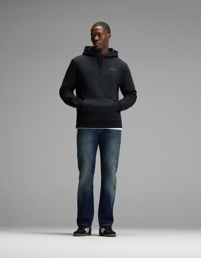 A man stands facing forward, wearing the Cozy Earth Men’s Cityscape Hoodie in black, paired with blue jeans, black slide sandals with white stripes, and a white shirt visible at the hem. The background is plain and gray.|Color:Jet Black