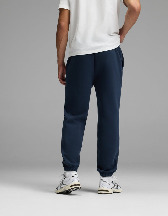 A person stands facing away, wearing a white T-shirt, Cozy Earth’s Men’s Cityscape Sweatpant in navy blue, and white sneakers. The background is plain and gray.|Color:Eclipse