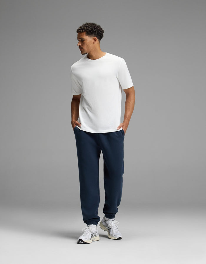 A man stands against a gray background, wearing a white t-shirt, white sneakers, and Cozy Earth’s Men’s Cityscape Sweatpant in navy blue, with his hands in his pockets and looking to the side.|Color:Eclipse