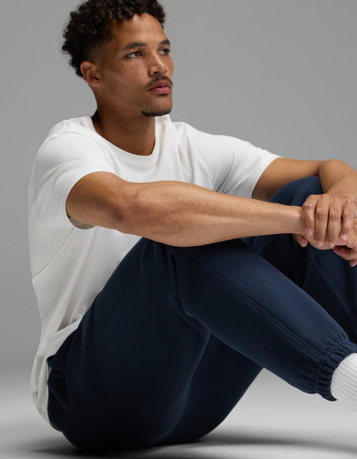 A man with short curly hair sits on the floor, arms resting on his knees, wearing a white t-shirt and Cozy Earth’s Men’s Cityscape Sweatpant in navy. He looks off to the side with a neutral expression against a plain gray background.|Color:Eclipse