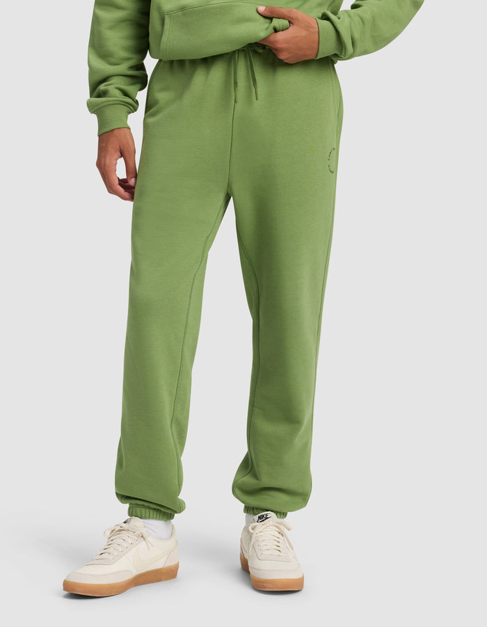 A person wears Cozy Earth's Men's Cityscape Sweatpant in green, paired with a matching hoodie and white-beige Nike sneakers, standing against a plain light background. Only the lower half of their body is visible.|Color:Clover