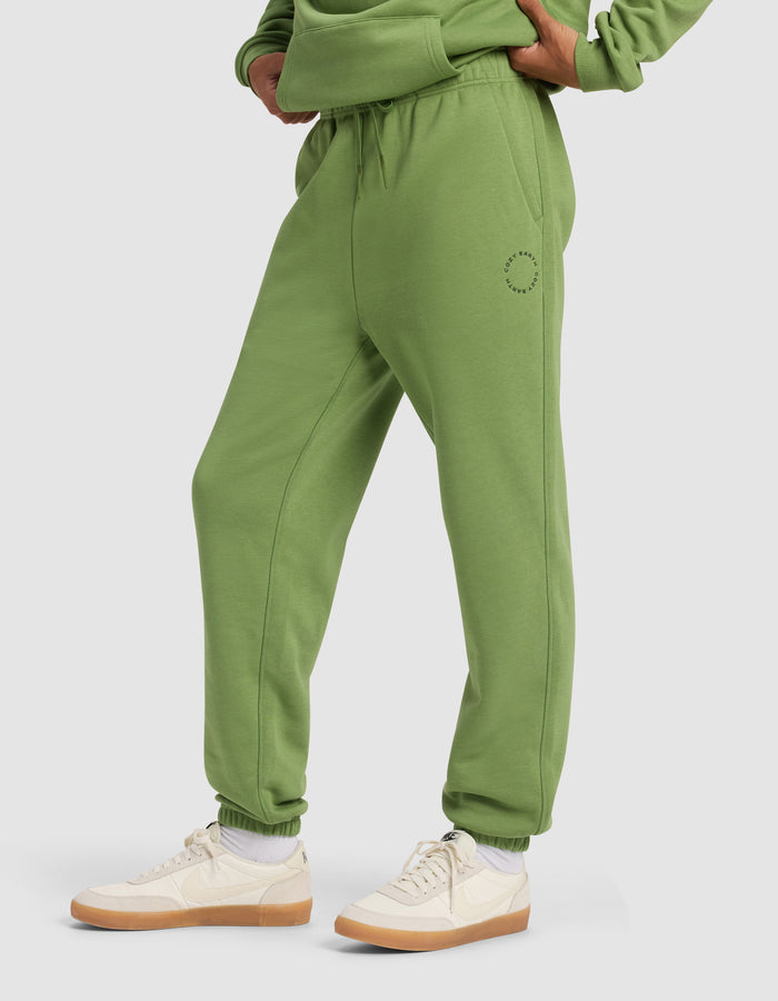A person wearing Cozy Earth Men's Cityscape Sweatpant in green with a matching sweatshirt, white socks, and beige sneakers stands hands on hips against a plain light background.|Color:Clover