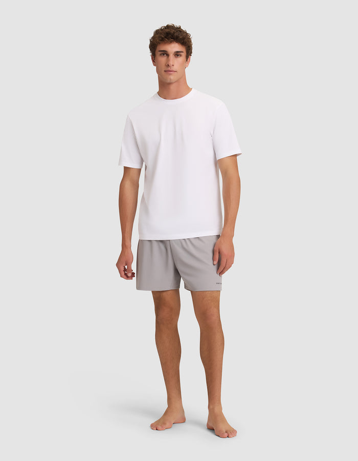 A young man stands barefoot against a light grey background, facing forward with a neutral expression. He is wearing Cozy Earth Men's Day & Night Shorts 5" in light grey and a plain white short-sleeve t-shirt. |Color:Stone