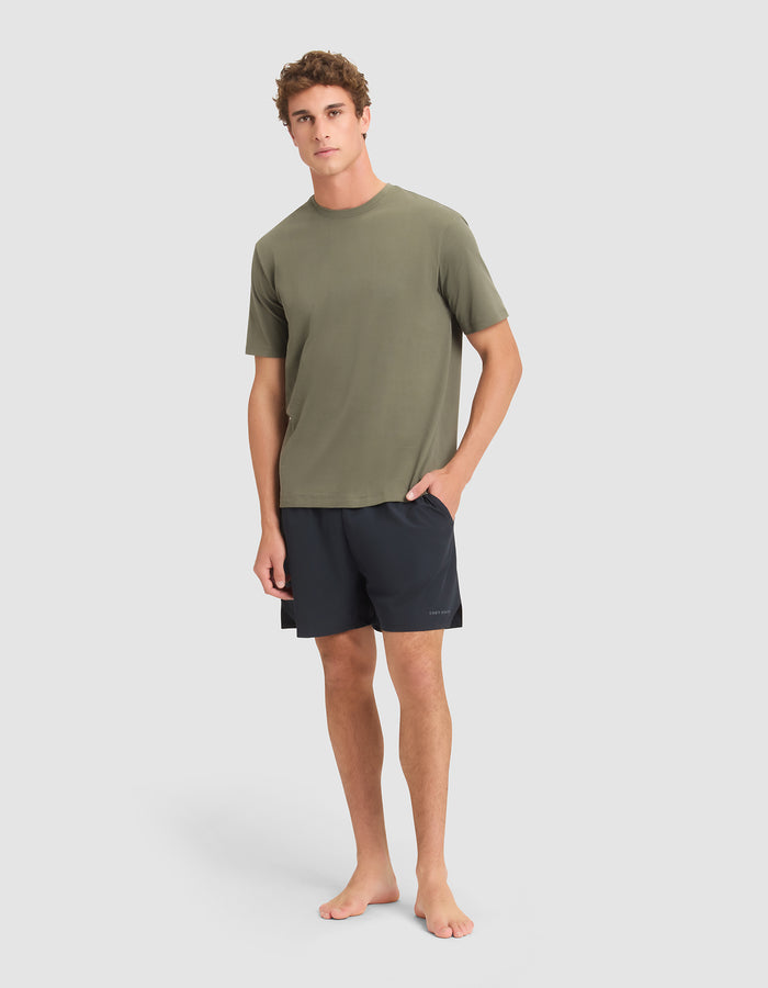 A young man stands barefoot against a plain background, wearing Cozy Earth Men's Day & Night Shorts 5" in black and an olive green short-sleeve t-shirt. His left hand is in his pocket, and he has a neutral expression. |Color:Jet Black