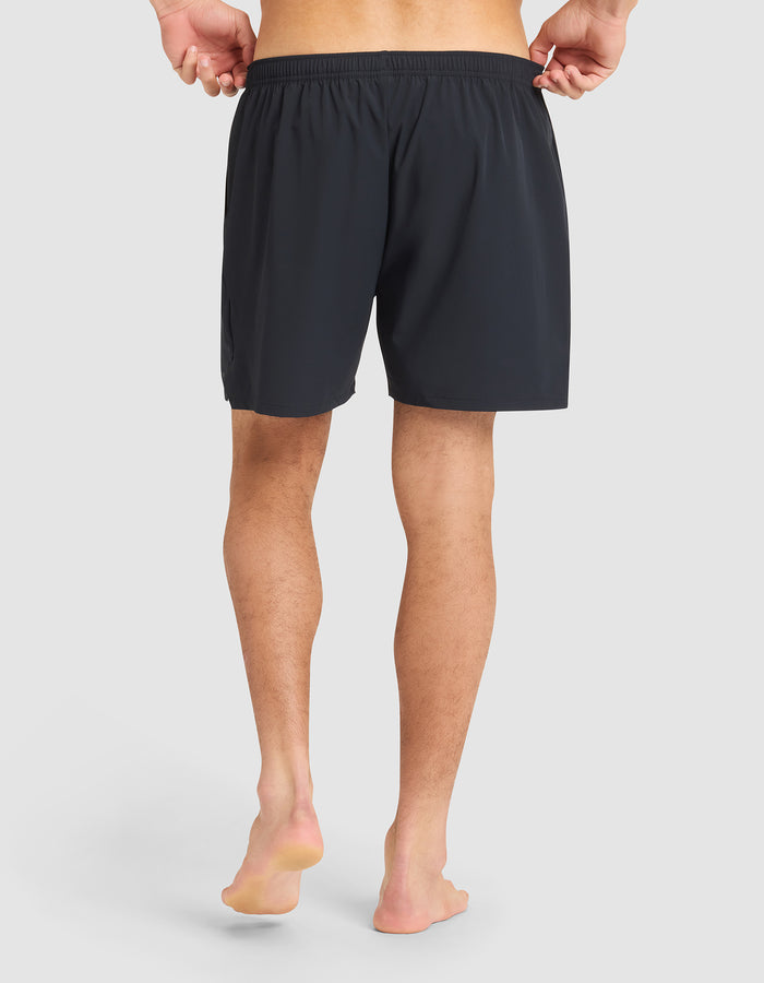 A barefoot man faces away from the camera, holding the waistband of Cozy Earth Men's Day & Night Shorts 5" in black against a plain light background. |Color:Jet Black