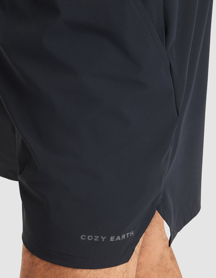 Close-up of a person wearing Cozy Earth Men's Day & Night Shorts 5" in black, with "COZY EARTH" printed in white near the bottom hem, standing against a plain light background. |Color:Jet Black