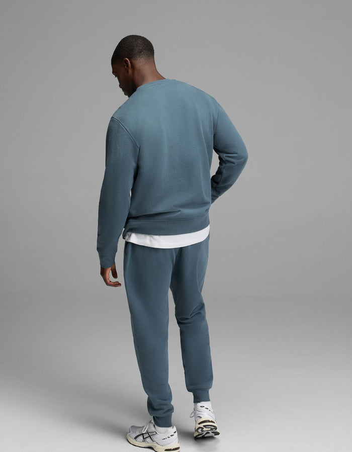 A person stands facing away against a plain gray background, wearing the Cozy Earth Men’s Dylan Crewneck & Jogger Set in blue-green over a white T-shirt and paired with white sneakers.|Color:Dawn