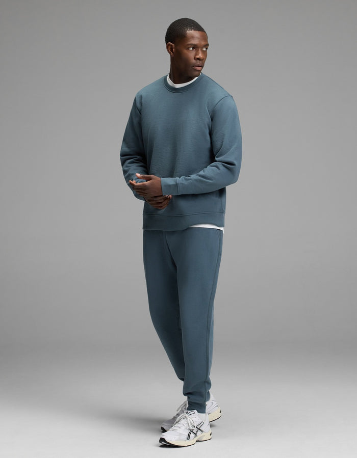 A man stands in a studio wearing the Cozy Earth Men’s Dylan Crewneck & Jogger Set paired with white sneakers. He looks to his left with his hands loosely clasped, against a plain light gray background.|Color:Dawn