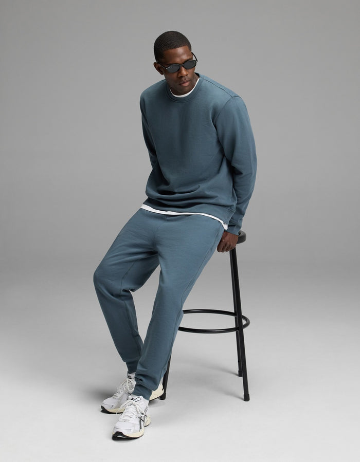 A person wearing the Men’s Dylan Crewneck & Jogger Set by Cozy Earth sits on a black stool against a plain gray background. They are also wearing sunglasses and white sneakers, and looking slightly to the side.|Color:Dawn