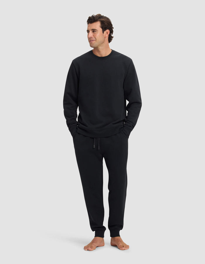 A man stands barefoot against a light gray background, wearing the Cozy Earth Men's Dylan Crewneck & Jogger Set in black. He has his hands in his pockets and gazes off to the side with a relaxed expression.|Color:Jet Black