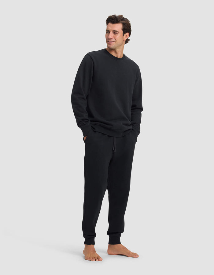 A man stands barefoot against a plain background, wearing the Cozy Earth Men's Dylan Crewneck & Jogger Set in black. He has his hands in his pockets and looks slightly to the side with a slight smile.|Color:Jet Black