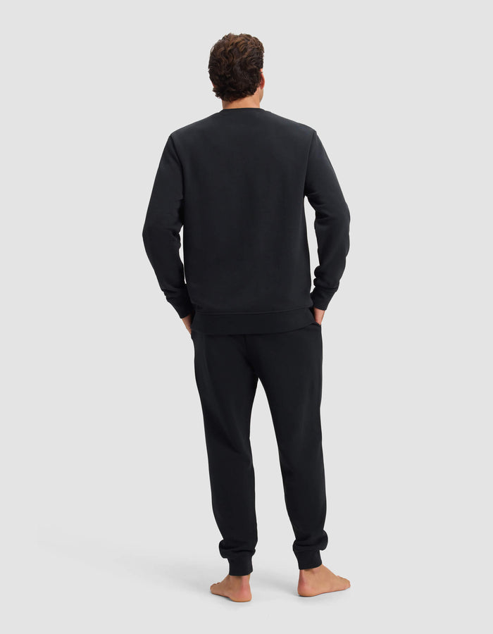 A person stands barefoot, facing away, wearing the Men's Dylan Crewneck & Jogger Set by Cozy Earth in black. Their hands are in their pockets against a plain light gray background.|Color:Jet Black