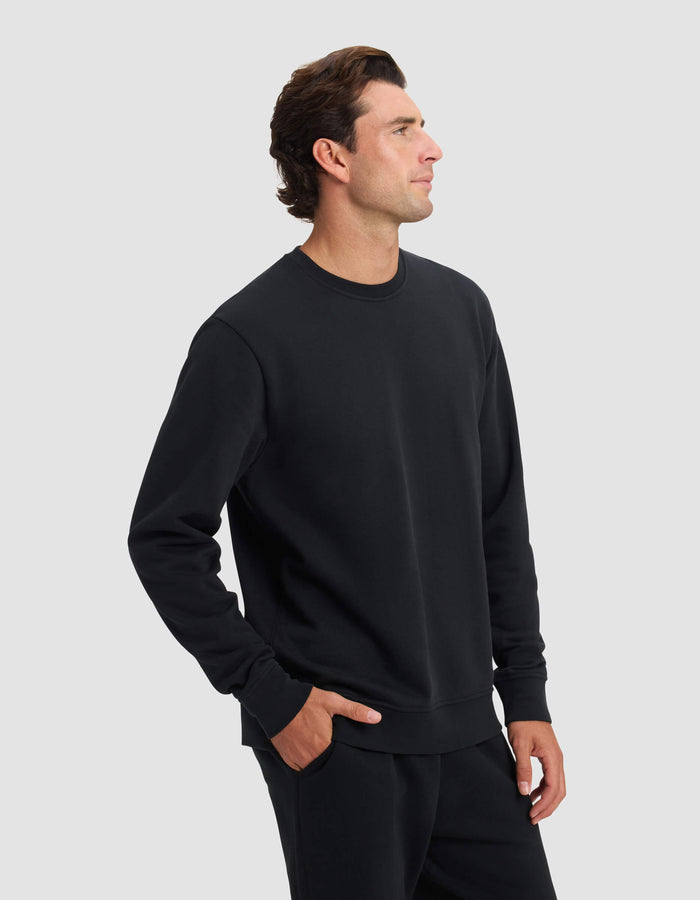 A man stands against a plain light gray background wearing the Cozy Earth Men's Dylan Crewneck and matching black pants, with one hand in his pocket and a neutral expression as he looks to his left.|Color:Jet Black