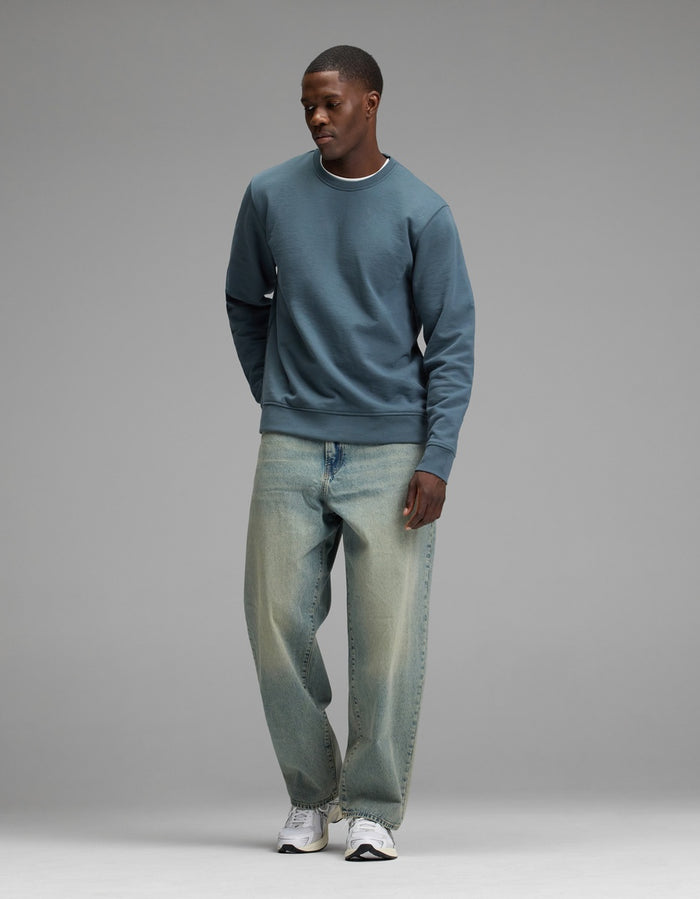 A person stands against a plain gray background wearing the Cozy Earth Men’s Dylan Crewneck, paired with loose light blue jeans and white sneakers, looking slightly downward with hands by their sides.|Color:Dawn