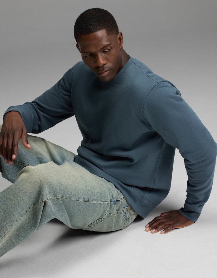 A man models the Cozy Earth Men’s Dylan Crewneck in blue, paired with light-wash jeans. He sits on the floor against a plain, neutral background, looking down with one hand on his knee and the other resting beside him.|Color:Dawn