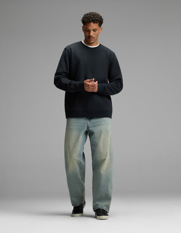 A man stands against a plain gray background, wearing the Cozy Earth Men’s Dylan Crewneck, paired with light blue relaxed-fit jeans and black sneakers. He looks down with his hands clasped together in front of him.|Color:Jet Black