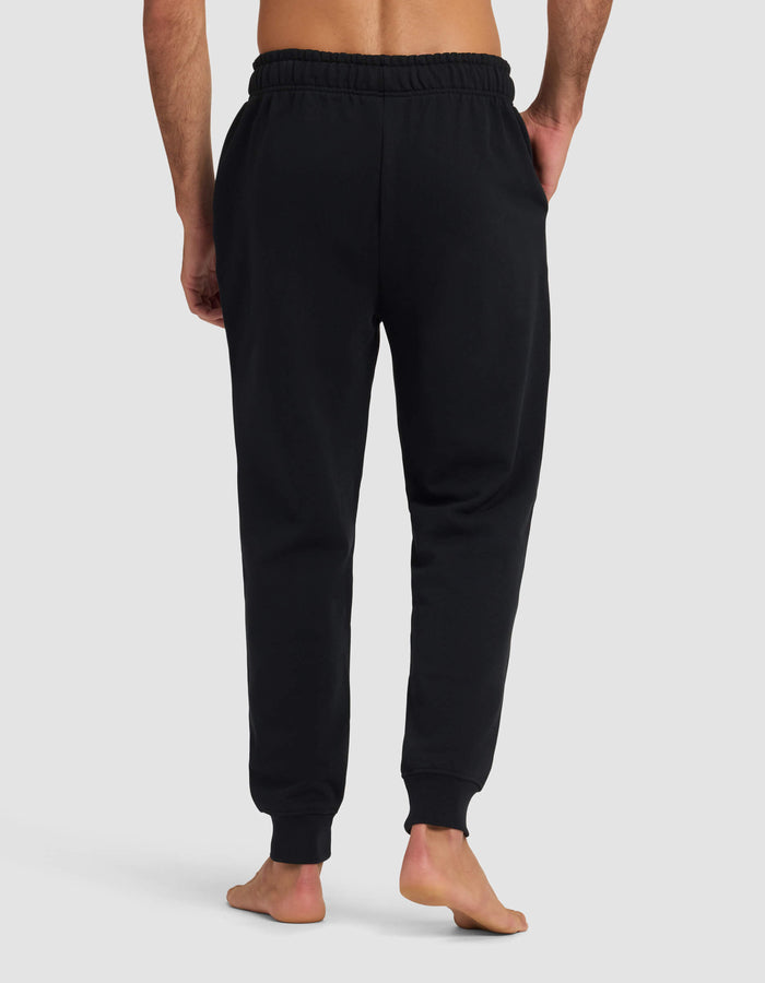 A barefoot man stands facing away, wearing Cozy Earth Men's Dylan Jogger—black sweatpants with an elastic waistband and cuffs—against a plain light gray background.|Color:Jet Black