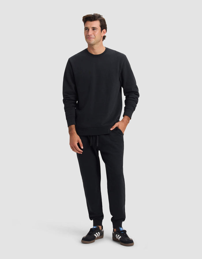 A man stands against a plain light gray background, wearing Cozy Earth's Men's Dylan Jogger in black with a matching sweatshirt and Adidas sneakers. He has short dark hair, a slight smile, and one hand in his pocket.|Color:Jet Black