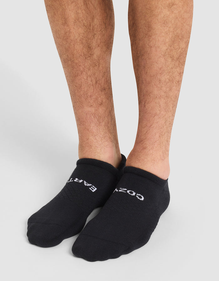 A person wearing Cozy Earth Essential Ankle Socks in black with white text stands on a light gray floor, showing their lower legs and feet partially covered by the socks. |Color:Black
