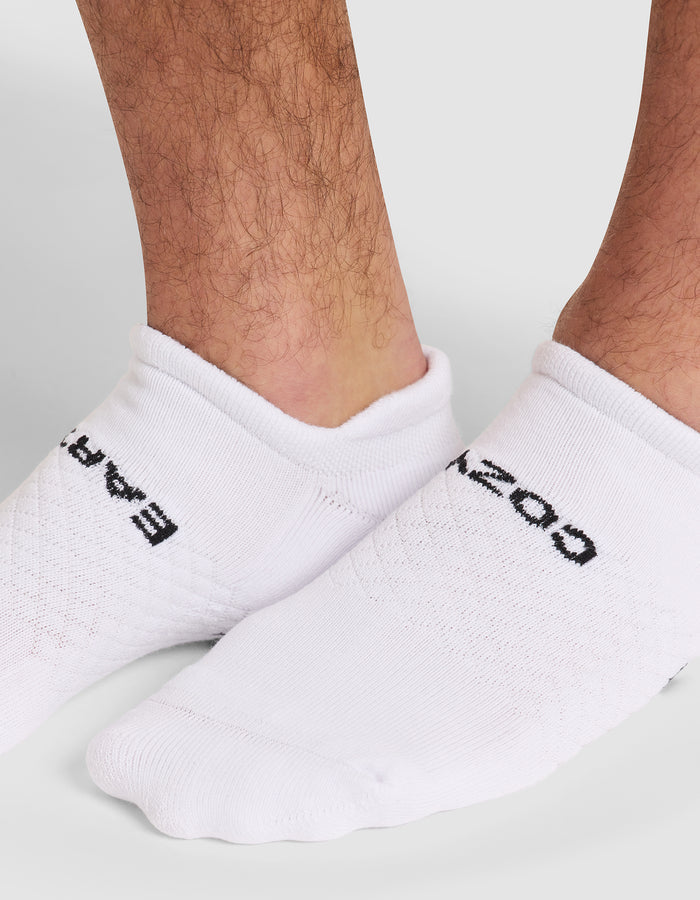 Close-up of a person's lower legs and feet wearing Cozy Earth's Essential Ankle Sock in white, featuring black text "EAR" on one sock and "OZ" on the other. The background is a plain light gray. |Color:White
