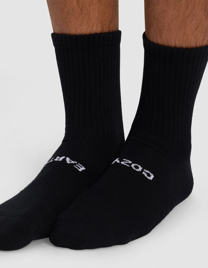 Close-up of lower legs and feet wearing Cozy Earth Essential Calf Socks in black. "EARTH" is printed on the left sock, "COSY" on the right. The plain, light background highlights the ribbed design.|Color:Black