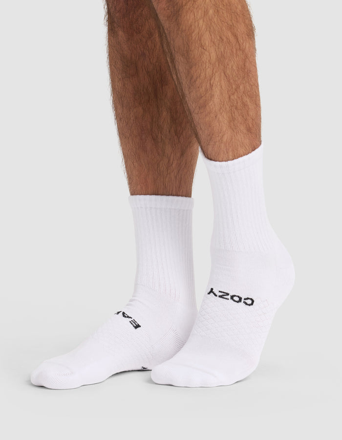 Someone wears Cozy Earth Essential Calf Sock 4-Pack—white crew socks with black text, covering ankles and lower legs—while standing on a light gray surface.|Color:White