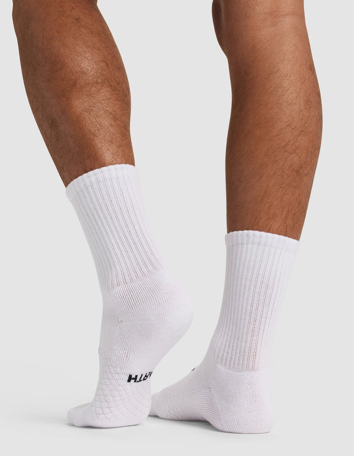 A person with medium skin tone stands on tiptoe, wearing Cozy Earth Essential Calf Socks in white. The ribbed crew socks feature a subtle texture and a small black logo near the sole, set against a plain white background.|Color:White