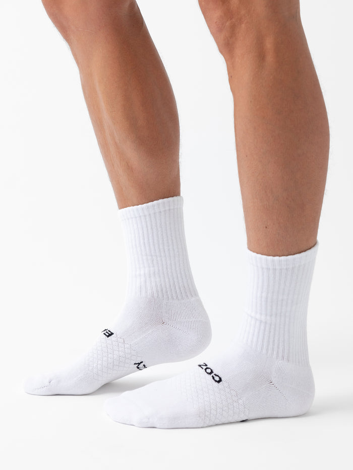 Two lower legs wearing Cozy Earth Essential Calf Socks stand on a white surface. The white socks feature ribbed cuffs, subtle textured patterns, and small black text near the toes. Sold as a 6-pack. |Color:White