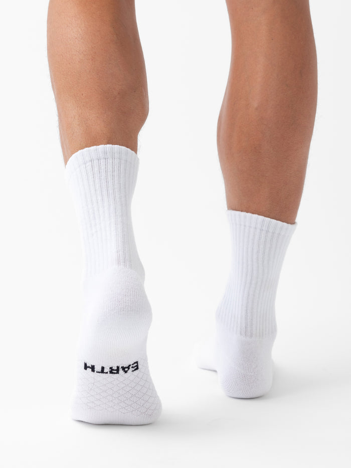 A person stands on tiptoes, viewed from behind, wearing white ribbed Essential Calf Socks by Cozy Earth. “EARTH” is printed in black on the sole of the left sock. The background is plain white. |Color:White