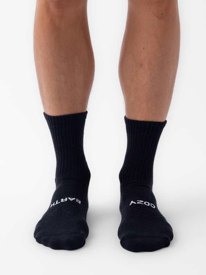 Someone wearing Cozy Earth Essential Calf Sock 6-Pack, black with white text at the top, stands on a white surface. The photo displays lower legs and feet from the front. |Color:Black