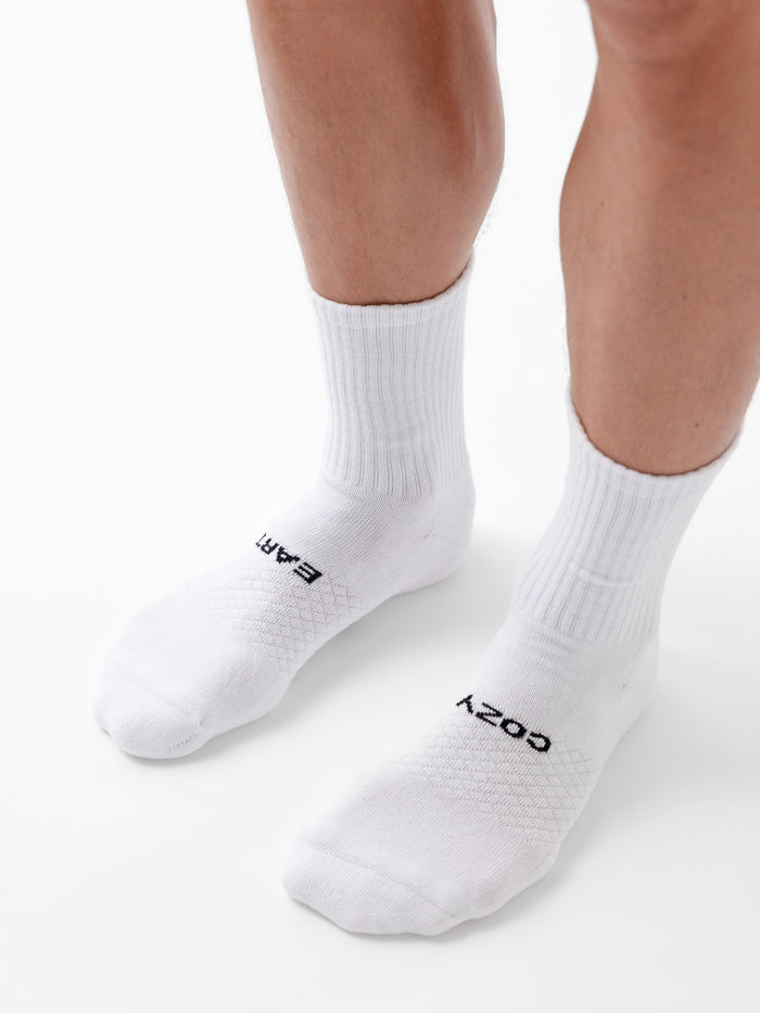 A person stands on a white surface wearing Cozy Earth’s Essential Calf Sock 8-Pack in white. The left sock reads "EARTH" and the right reads "COZY" in black letters near the toes. |Color:White