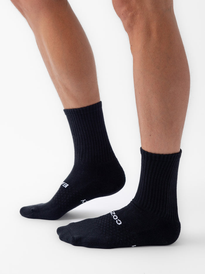 A person stands on a white background wearing Cozy Earth’s Essential Calf Sock (8-Pack) in black, with "COZY" visible on the side of one sock and one foot slightly in front of the other. |Color:Black