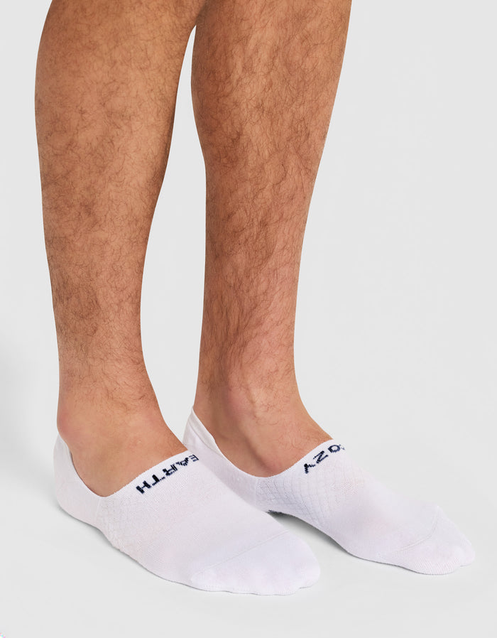 A person wears Cozy Earth Men's Essential No-Show Socks in white with dark text near the toes, standing on a light gray surface and displaying their lower legs and feet.