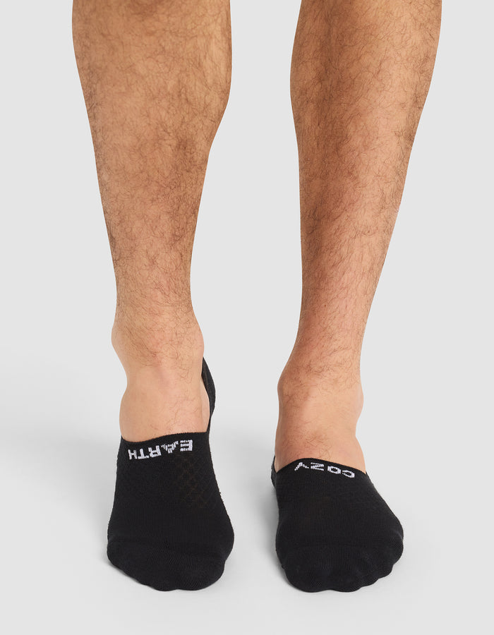 A person with bare legs wears Cozy Earth Essential No-Show Socks in black, featuring "EARTH" on the left and "COZY" on the right in white text. The background is plain and light-colored. |Color:Black