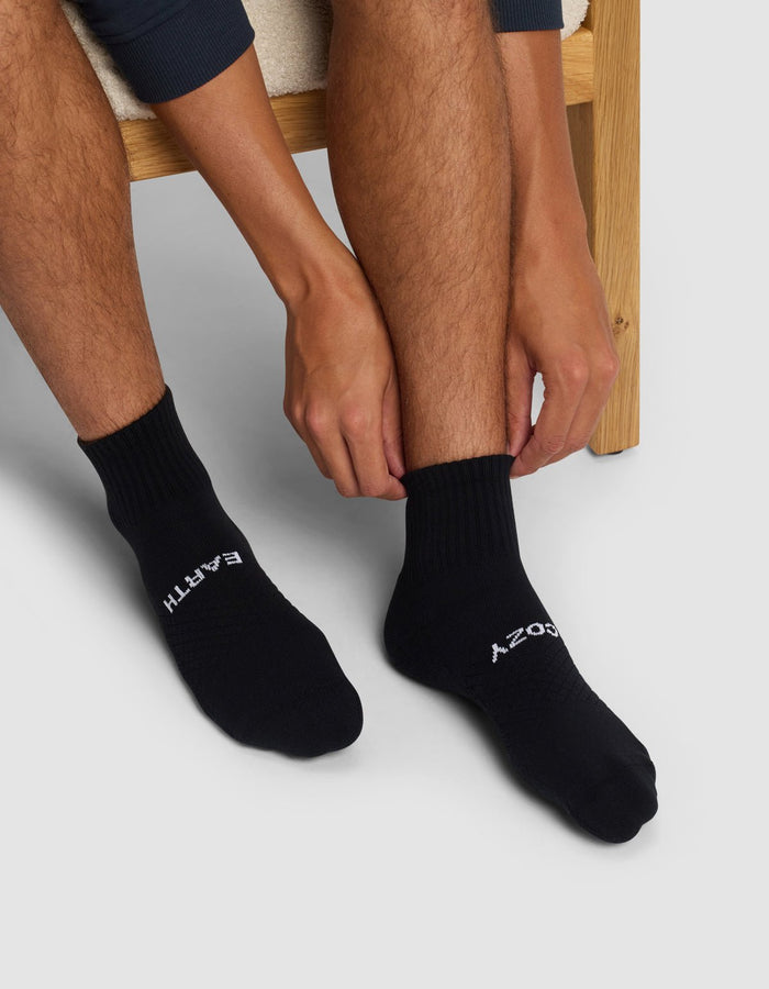 Seated on a wooden chair, a person wearing Cozy Earth Essential Quarter Socks with "EARTH" on the left and "SOCKS" on the right adjusts the right sock with both hands.|Color:Black