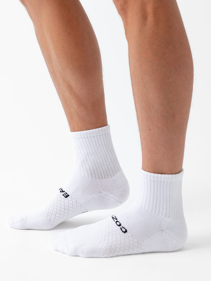 A person wearing Cozy Earth Essential Quarter Sock 6-Pack, featuring white socks with black text on top, stands against a white background. Only their lower legs and feet are visible. |Color:White